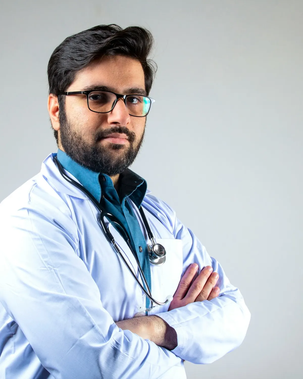 A doctor in a white coat and glasses, arms folded, against a plain background